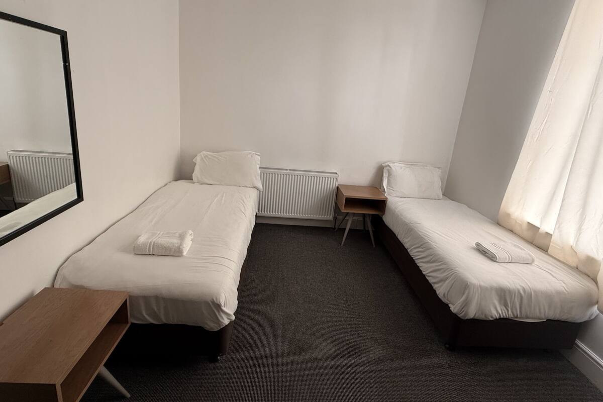 A hotel room with two single beds, a small wooden table, a radiator, and a window with curtains.