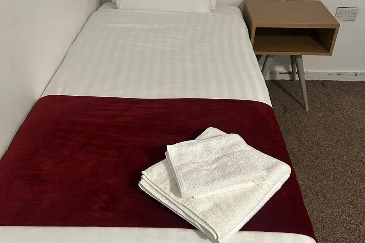 A single-bed room with a red blanket, white pillows, and a small wooden side table.