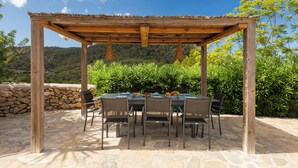 A covered outdoor dining area with a stone floor, wooden pergola, and a view of greenery and hills in the background.