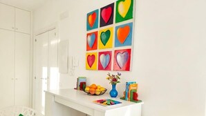 Ein weißes Zimmer mit einem Schreibtisch, einem Stuhl, einem Regal mit Büchern und einer Vase, sowie einer Wand mit herzförmigen Bildern.
