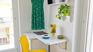 Ein kleines Zimmer mit einem weißen Schreibtisch, einem gelben Stuhl, einem grünen Kleid an einem Kleiderbügel, einem an der Wand montierten Fernseher und einer Zimmerpflanze.