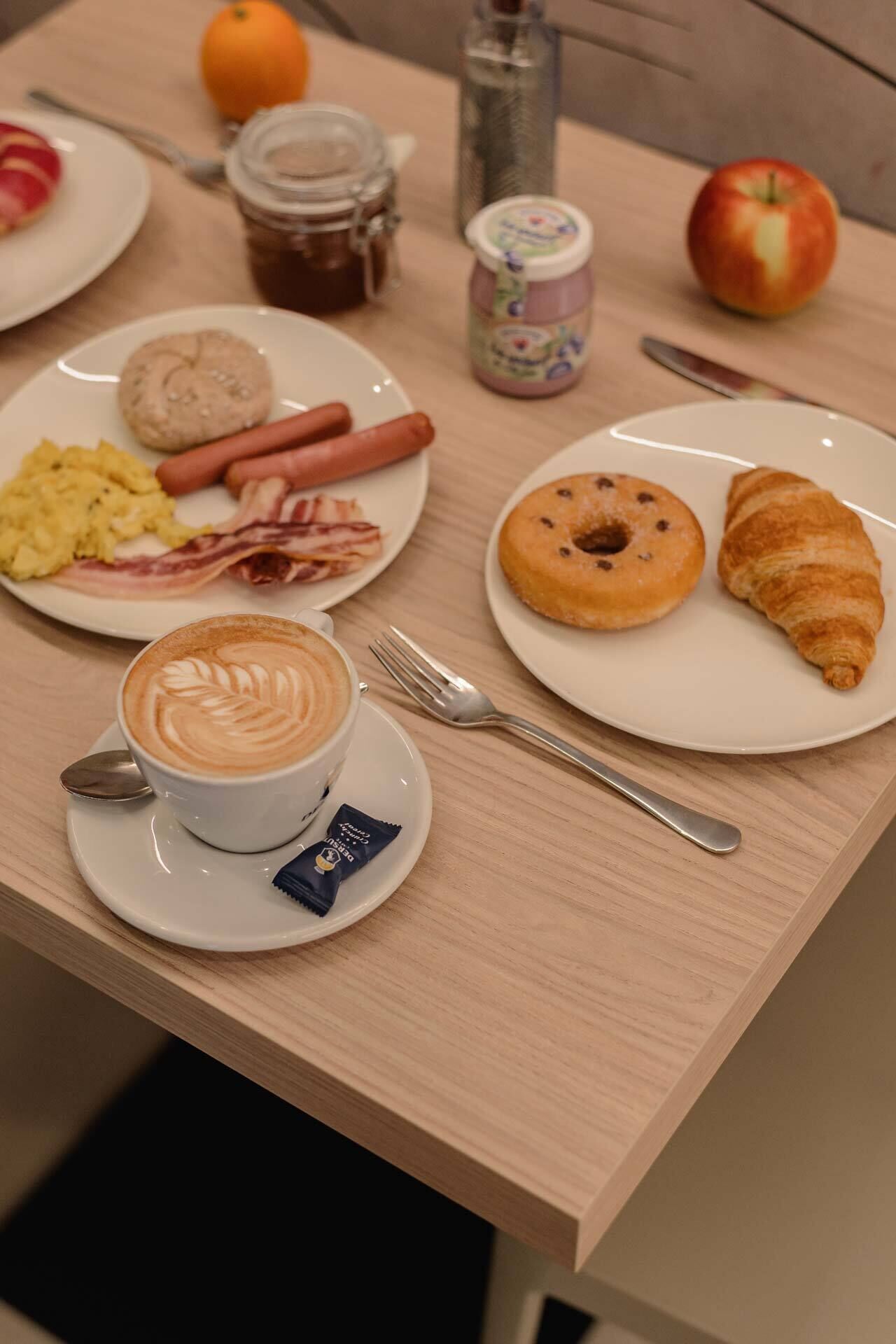 Ein Frühstückstisch mit einem Teller Eier, Speck, Würstchen, einem Croissant, einem Donut und einer Kaffeetasse mit Latte-Art.