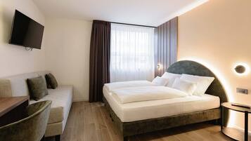 A hotel room with a bed, a sofa, a TV mounted on the wall, and a window with curtains.