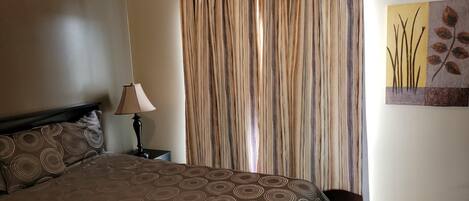 A bedroom with a bed, a nightstand, a lamp, a curtain, and a wall art piece.