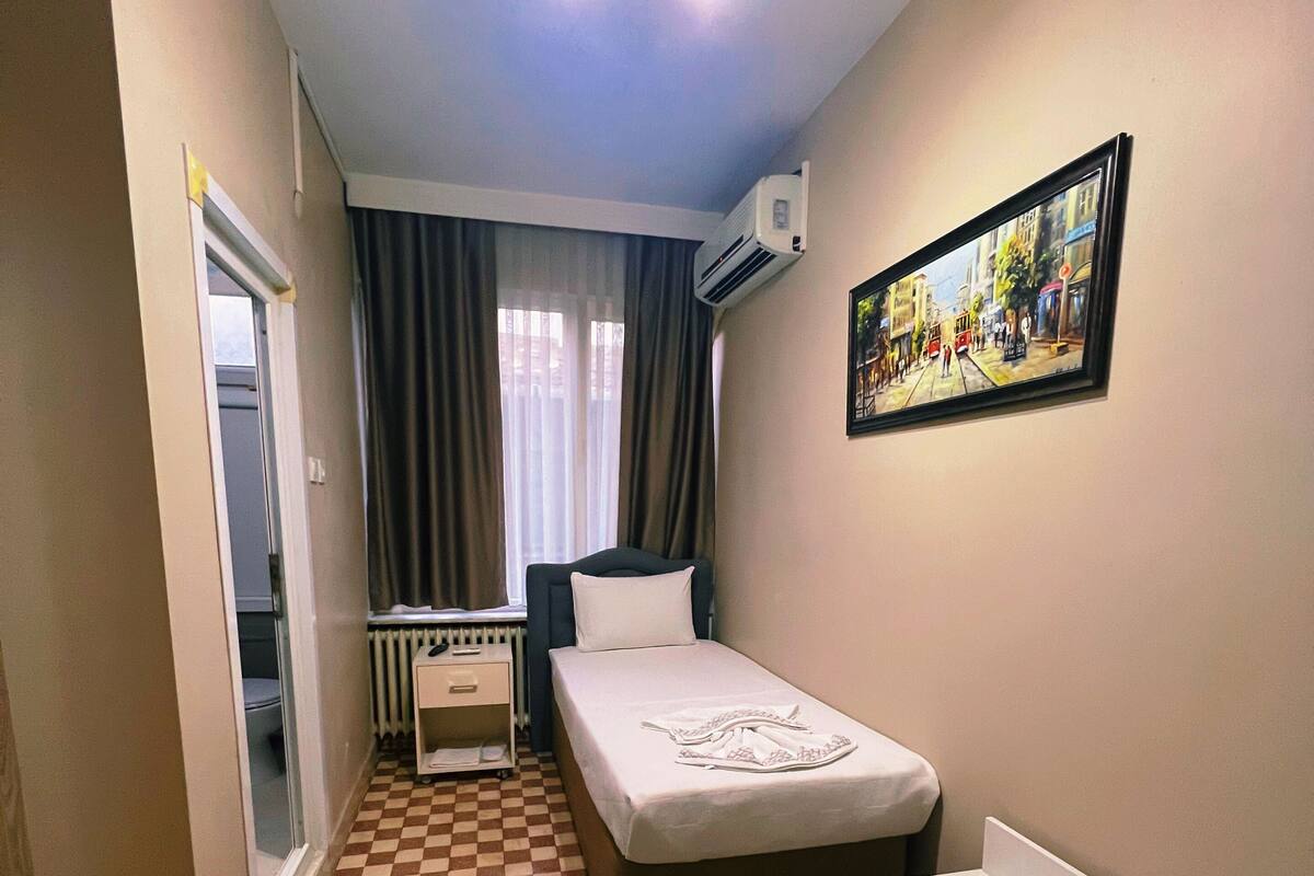 A compact hotel room with a checkered floor, a bed, a small bedside table, a white bathtub, a radiator, and a painting on the wall.