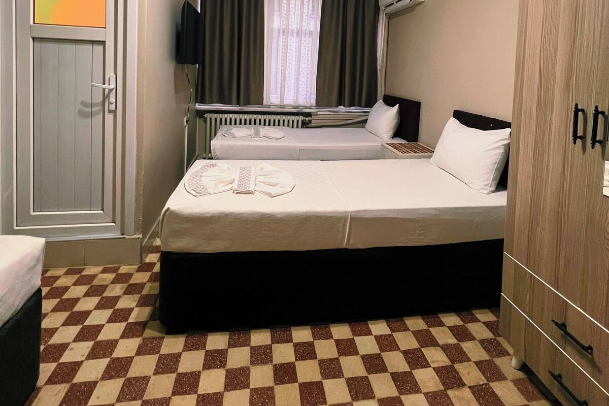 A hotel room with two beds, a checkered floor, a radiator, a window with curtains, and a wardrobe.