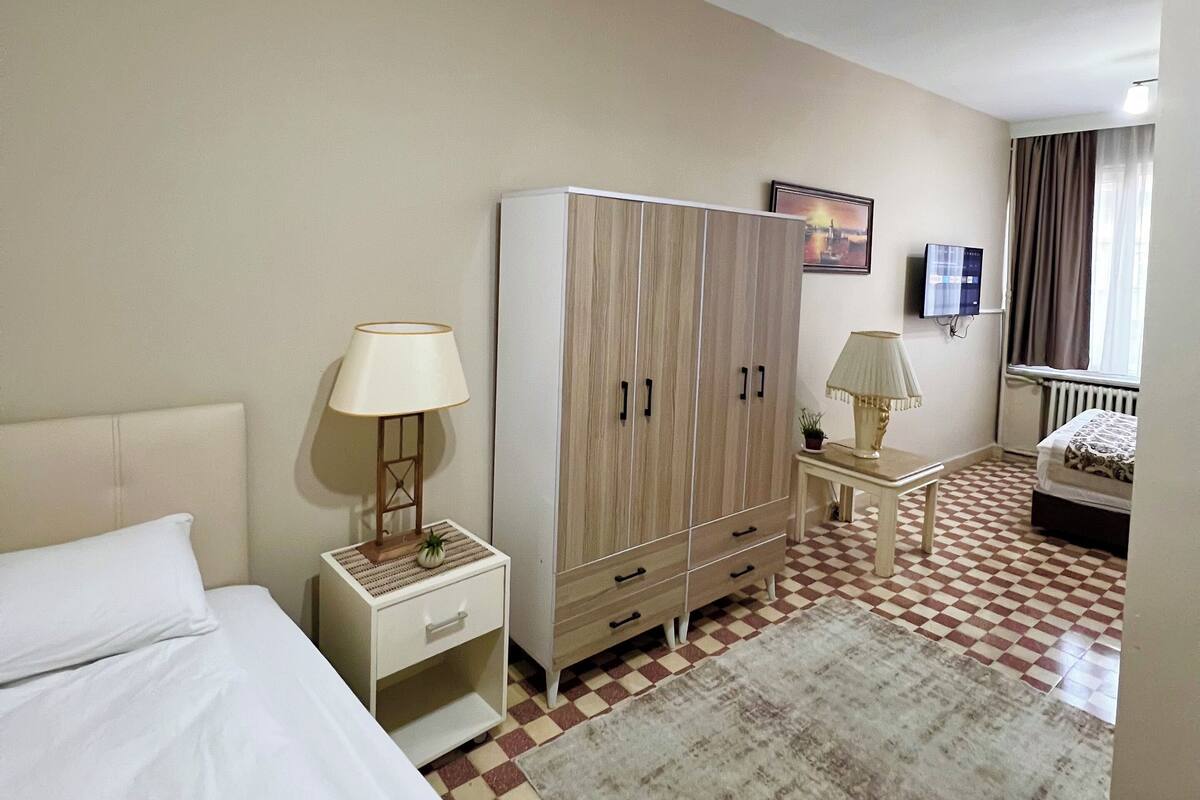A hotel room with a bed, a wardrobe, a nightstand, a lamp, a checkered floor, and a window with curtains.