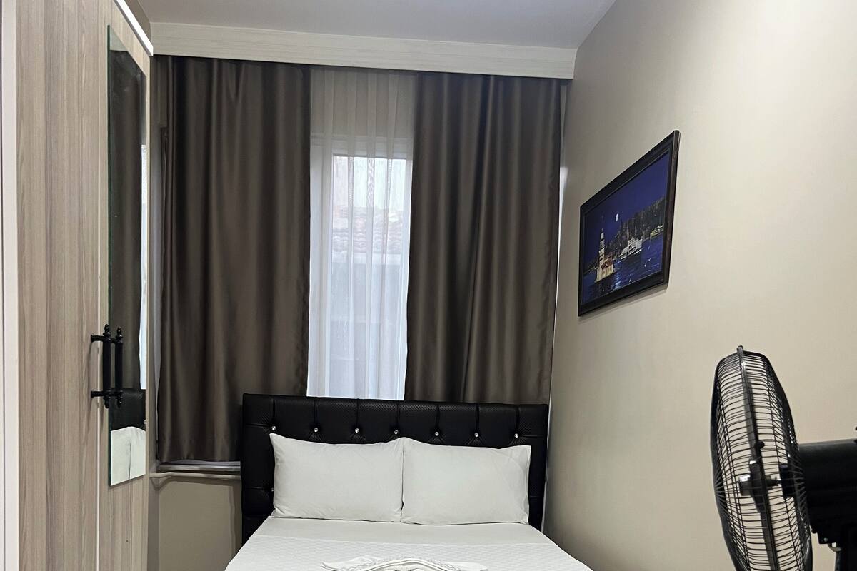 A hotel room with a bed, a desk, a chair, a fan, and a checkered floor.
