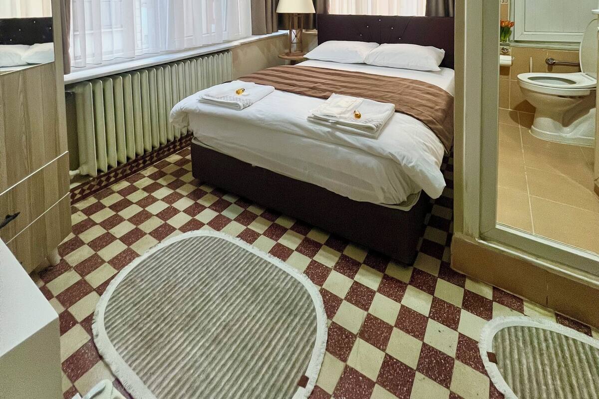 A hotel room with a checkered floor, a bed with white linens, a bedside lamp, a window with curtains, and a radiator.