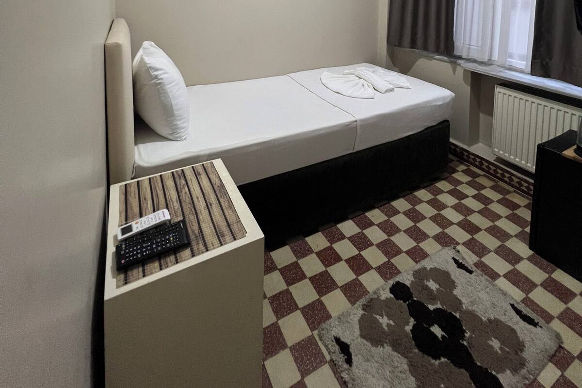 A small, single-bed room with a checkered floor, a remote control, and a window with curtains.