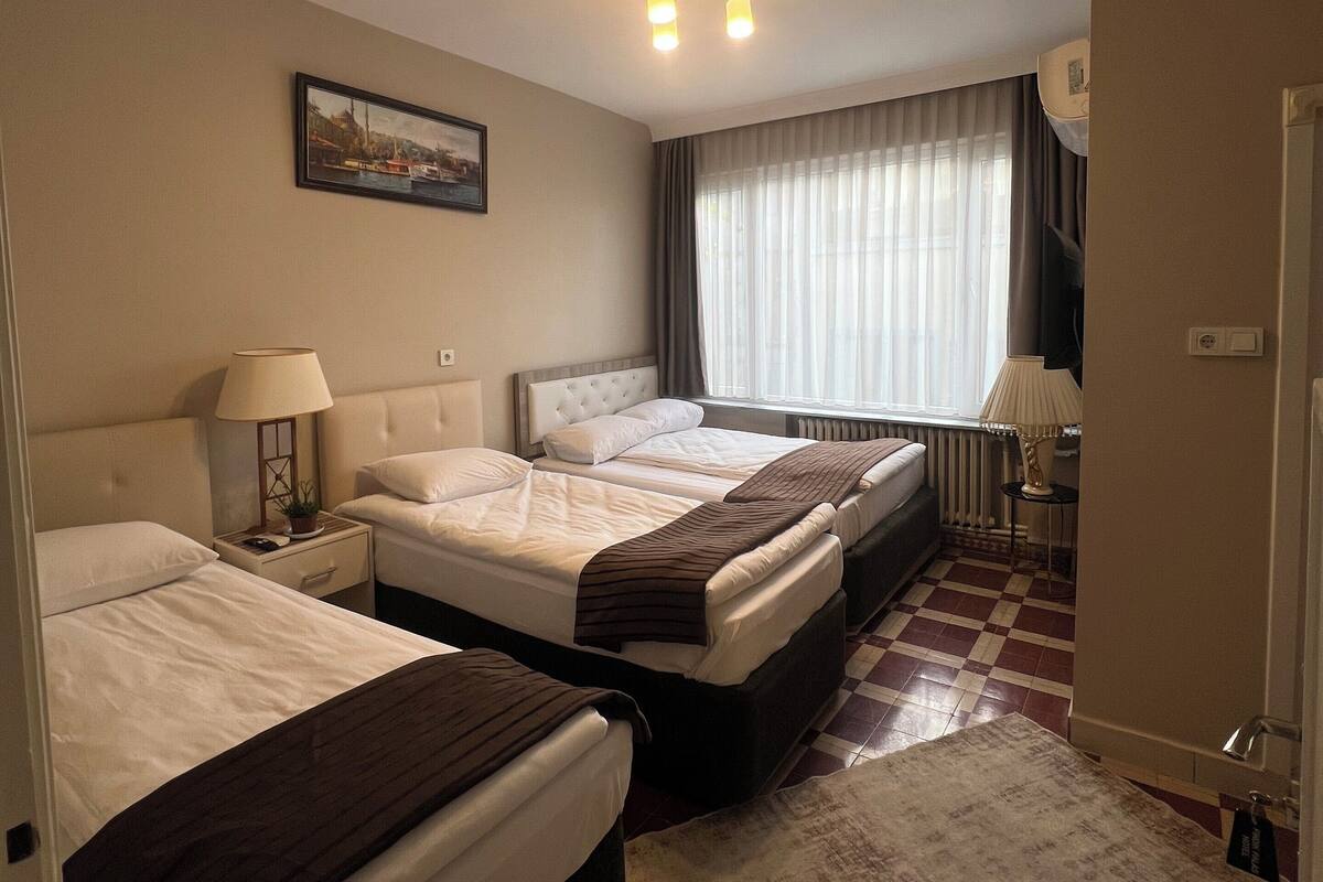 A hotel room with three beds, a large window with curtains, a ceiling light fixture, and a painting on the wall.