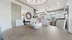 A modern dining room with a round wooden table, striped chairs, a chandelier, and a view into the kitchen.
