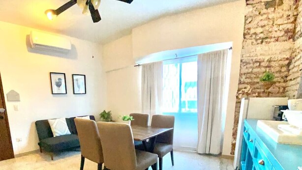 A dining area with a table and chairs, a ceiling fan, a stone wall, and a window with curtains. - Furlong1 Apartment in down Town city near the malecón and restaurants (La Paz)