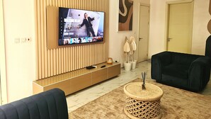 Un salon moderne avec un téléviseur à écran plat fixé sur un mur recouvert de panneaux en bois, un canapé foncé, une table basse ronde en bois et une porte visible au fond.