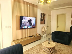 A modern living room with a flat-screen TV mounted on a wooden panel wall, a dark sofa, a round wooden coffee table, and a door in the background. - Lux 3BD/3BA Flat | Parkview Ikoyi | NG01 (Lagos)