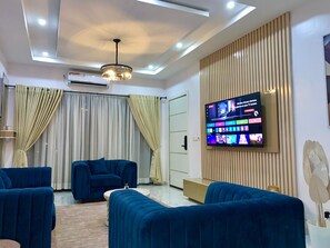 A modern living room with blue sofas, a flat-screen TV mounted on a wooden panel, and a ceiling light fixture. - Lux 3BD/3BA Flat | Parkview Ikoyi | NG01 (Lagos)