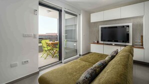 Een moderne woonkamer met een bank, een flatscreen televisie en een schuifpui naar een balkon met een tafel en stoelen.