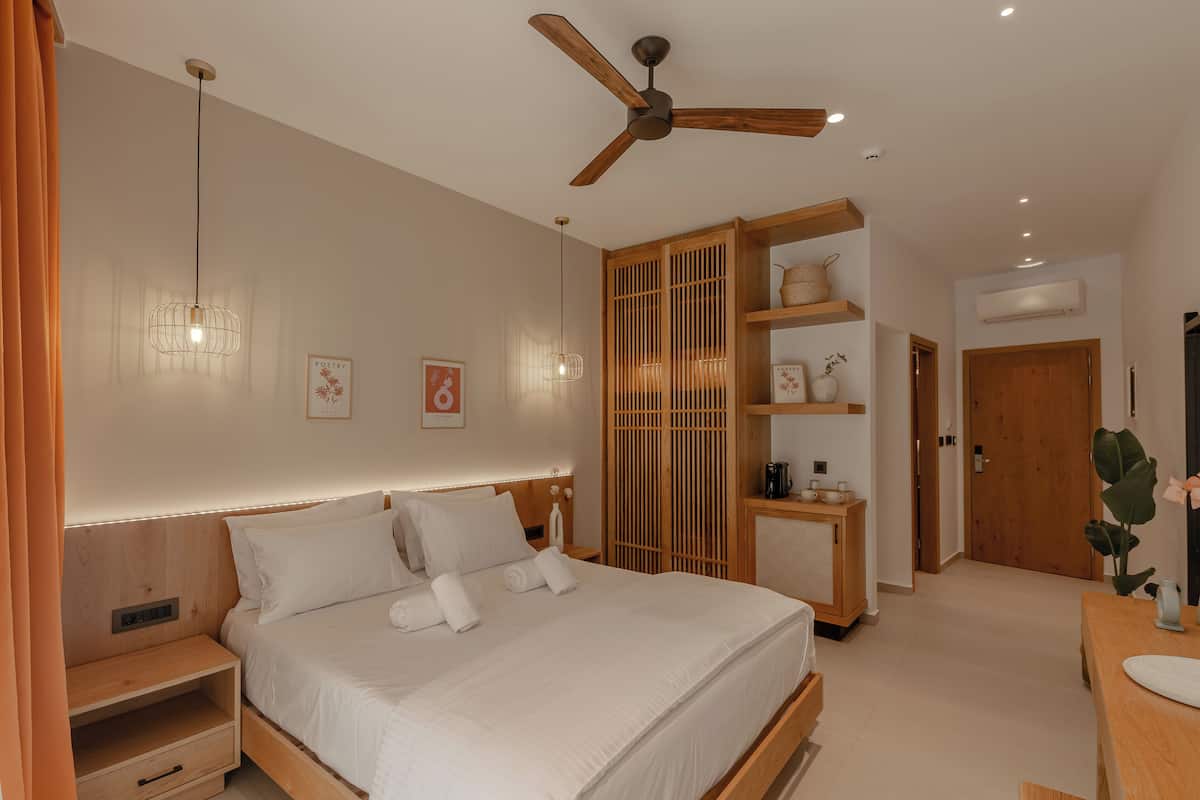 A modern hotel room with a large bed, a ceiling fan, a wooden wardrobe, and a bedside table with a lamp.