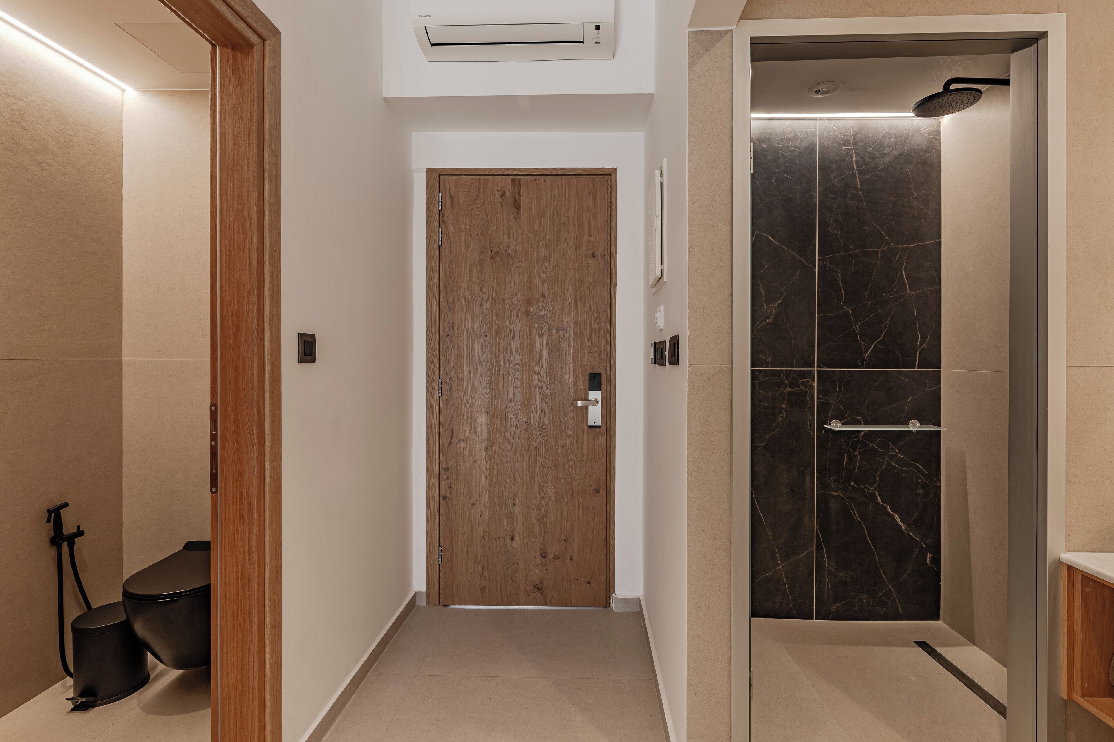 Un pasillo con puerta de madera, zona de ducha con azulejos de mármol y un inodoro negro.