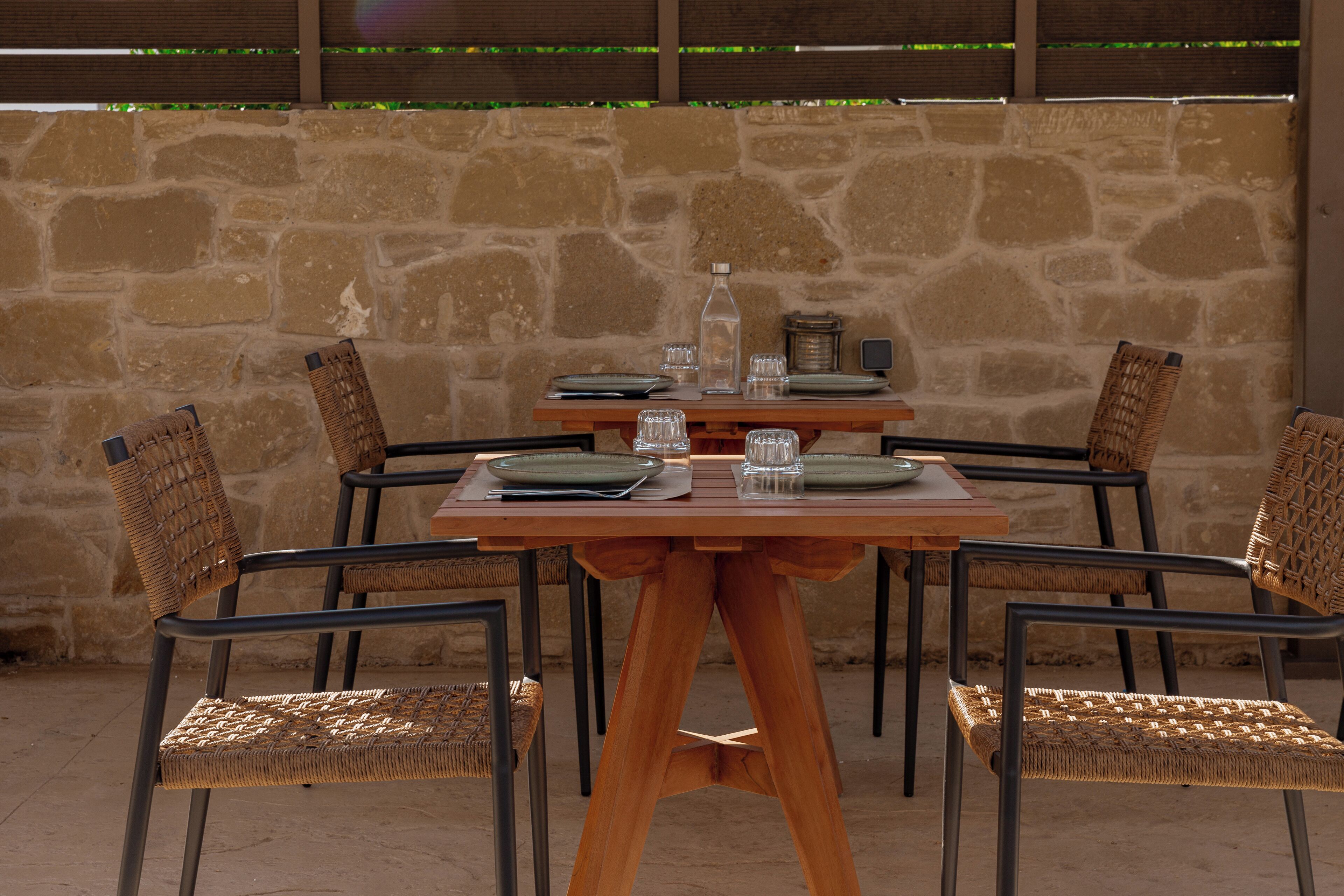 A wooden table with four wicker chairs, set against a stone wall.