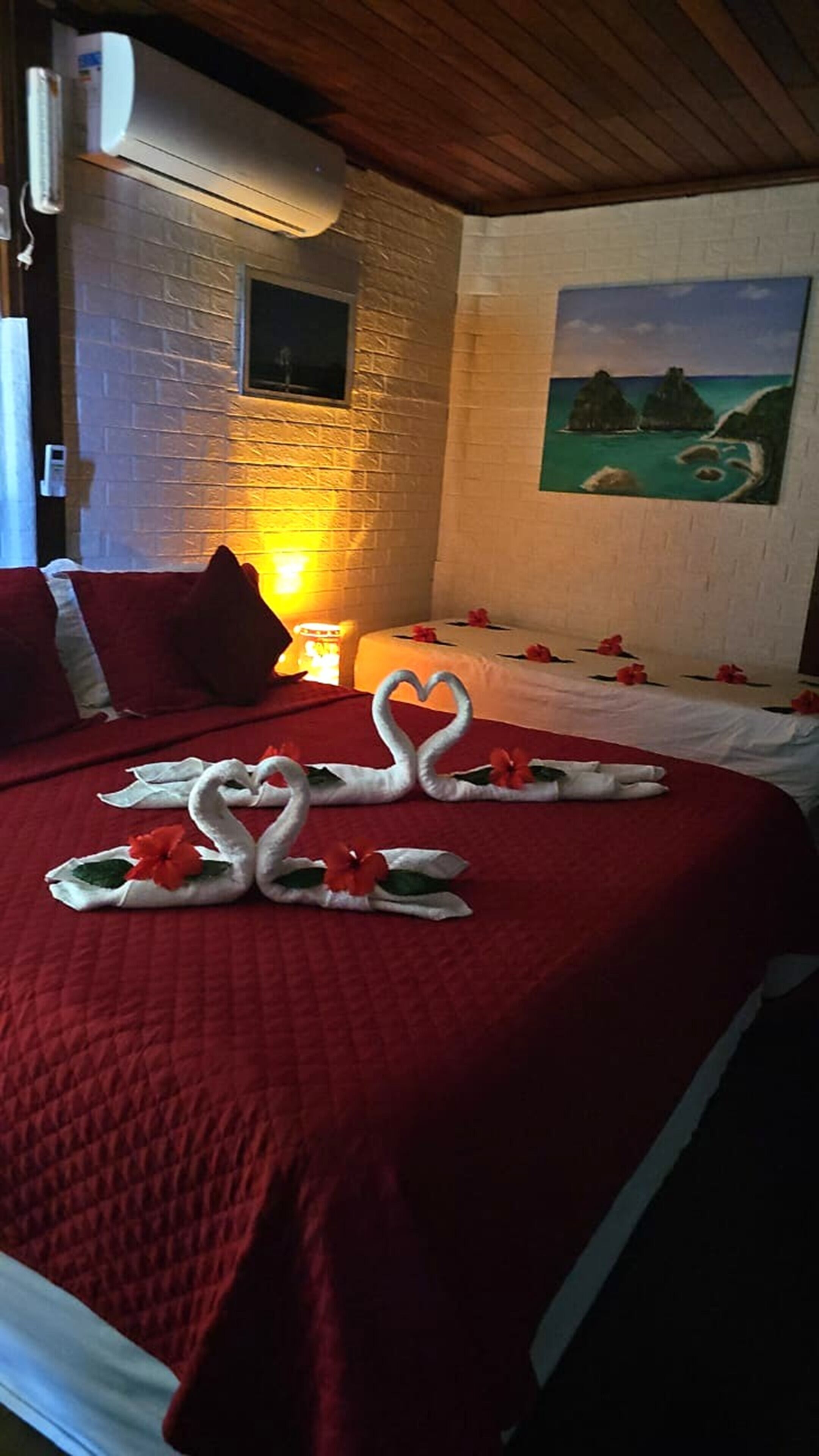A hotel room with a red quilted bedspread, a painting of a beach scene, and a wall-mounted air conditioner.