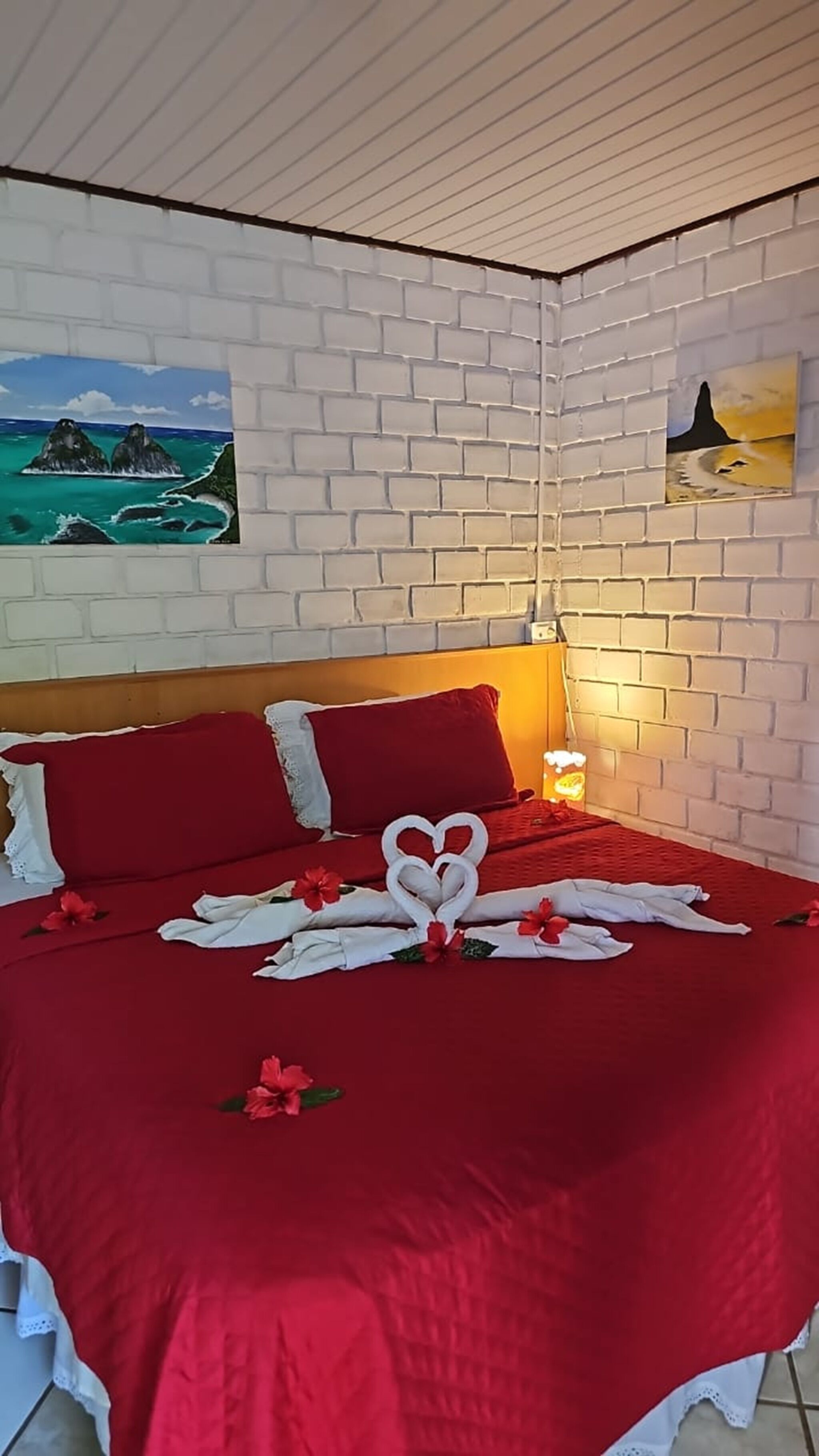 A bedroom with a brick-patterned wall, a bed with red bedding, and two paintings.