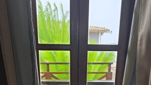 A room with a view through a window with wooden shutters, a tiled floor, and a view of a balcony with a wooden railing and green plants. - Pousada Capim Açu (Fernando de Noronha)