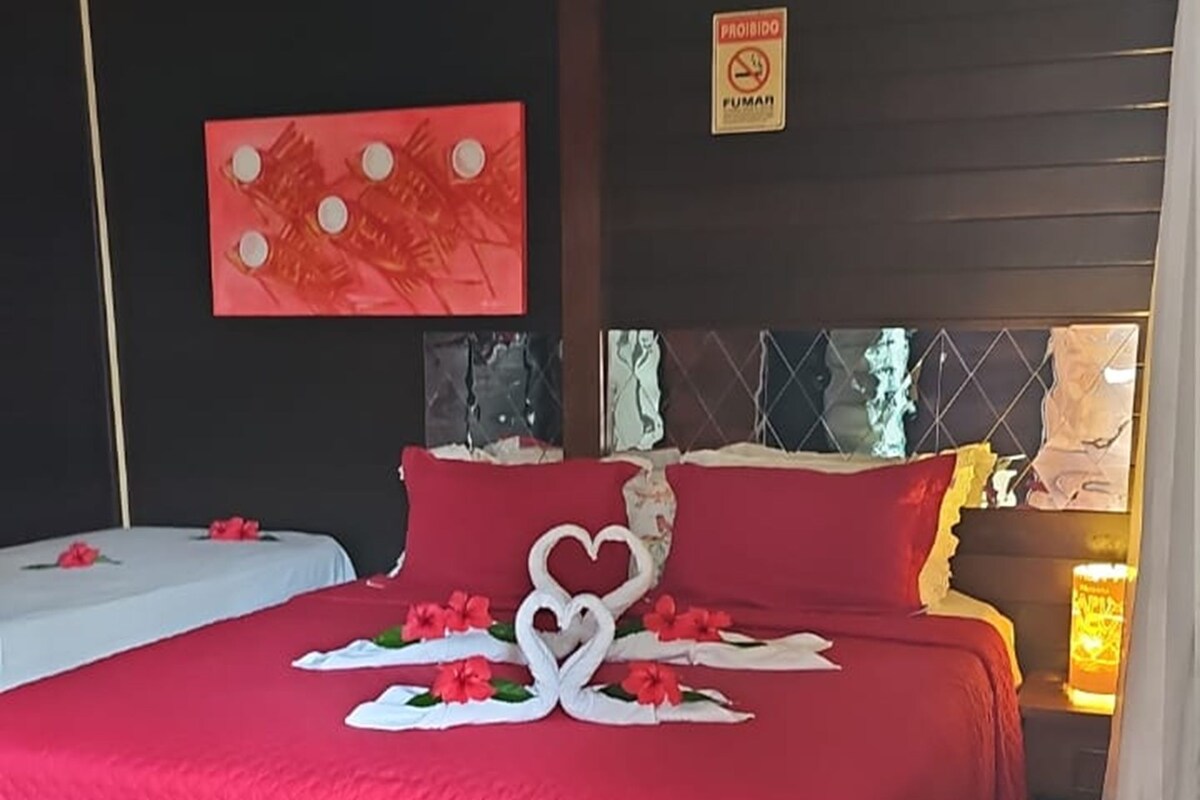 Une chambre d’hôtel avec deux lits, une couvre-lit rouge et blanche, une décoration en forme de cœur faite avec une serviette, et un panneau indiquant « Fumer interdit » accroché au mur.