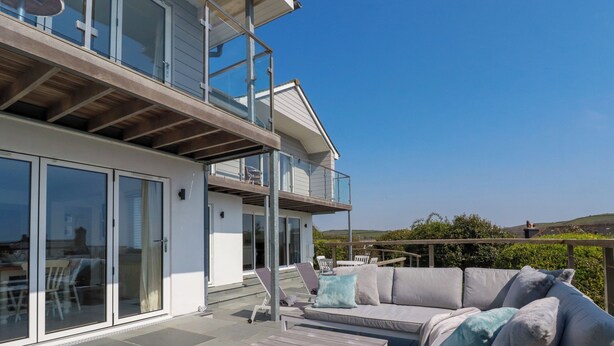 Un patio moderno al aire libre con un sofá gris, una mesa de madera y un ventanal de vidrio que da a un balcón.