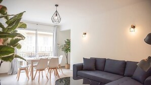 A modern living room with a grey sofa, a glass coffee table, and a dining area with white chairs and a table. There are two pendant lights and wall sconces. - Comfortable apartment in the heart of Antibes (Antibes)