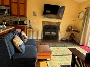 A living room with a sofa, ottoman, flat-screen TV, fireplace, kitchen area, and a clock on the wall.