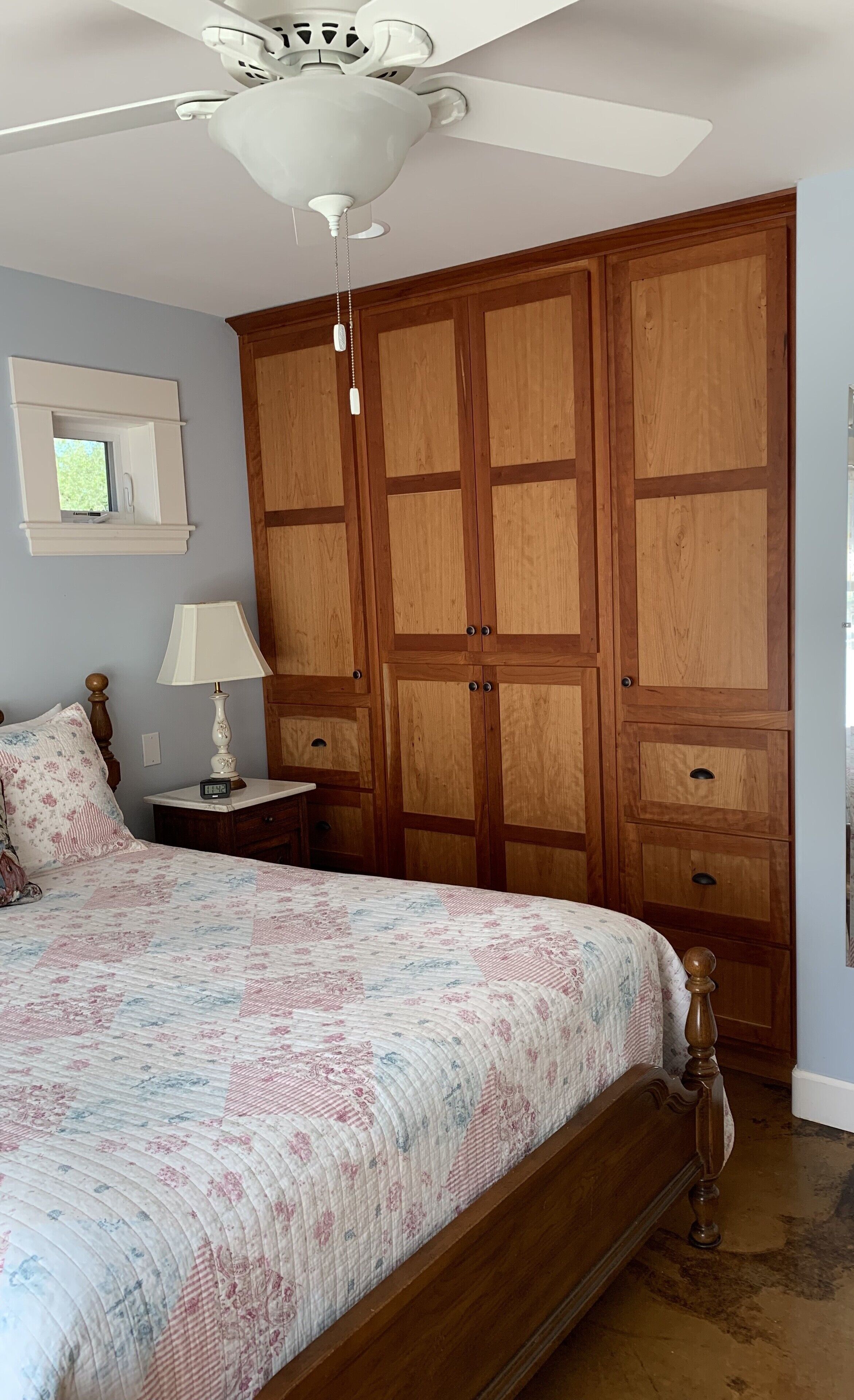A bedroom with a large bed, wooden wardrobe, ceiling fan, bedside lamp, and a small table with a clock.