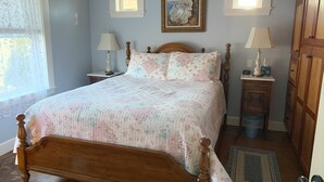 Un dormitorio con una cama de madera, dos mesitas de noche con lámparas, un ventanal con cortinas, un cuadro en la pared y un armario de madera.