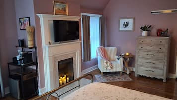 Ein gemütliches Zimmer mit Kamin, Fernseher, Sessel, Kommode und einem Fenster mit Vorhängen.