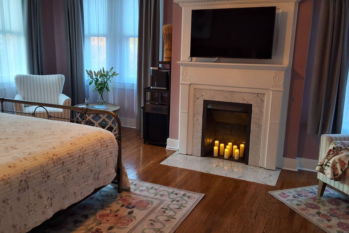 A hotel room with a bed, a fireplace with lit candles, a television mounted above the fireplace, a chair, and a small table with a vase of flowers.
