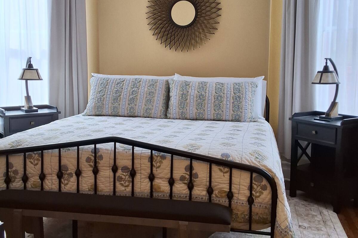 A bedroom with a bed, two bedside tables, a wall-mounted mirror, and a window with curtains.