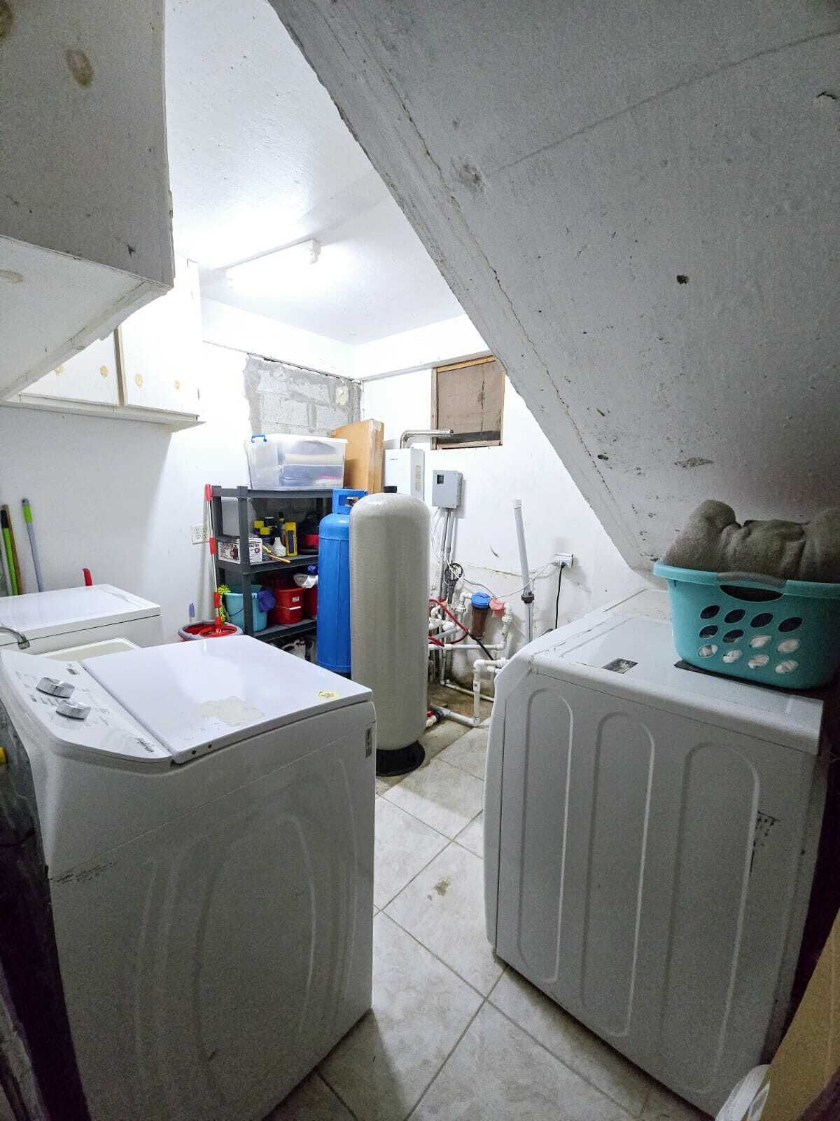 Een kleine wasruimte met twee wasmachines, een boiler en diverse schoonmaakmiddelen.