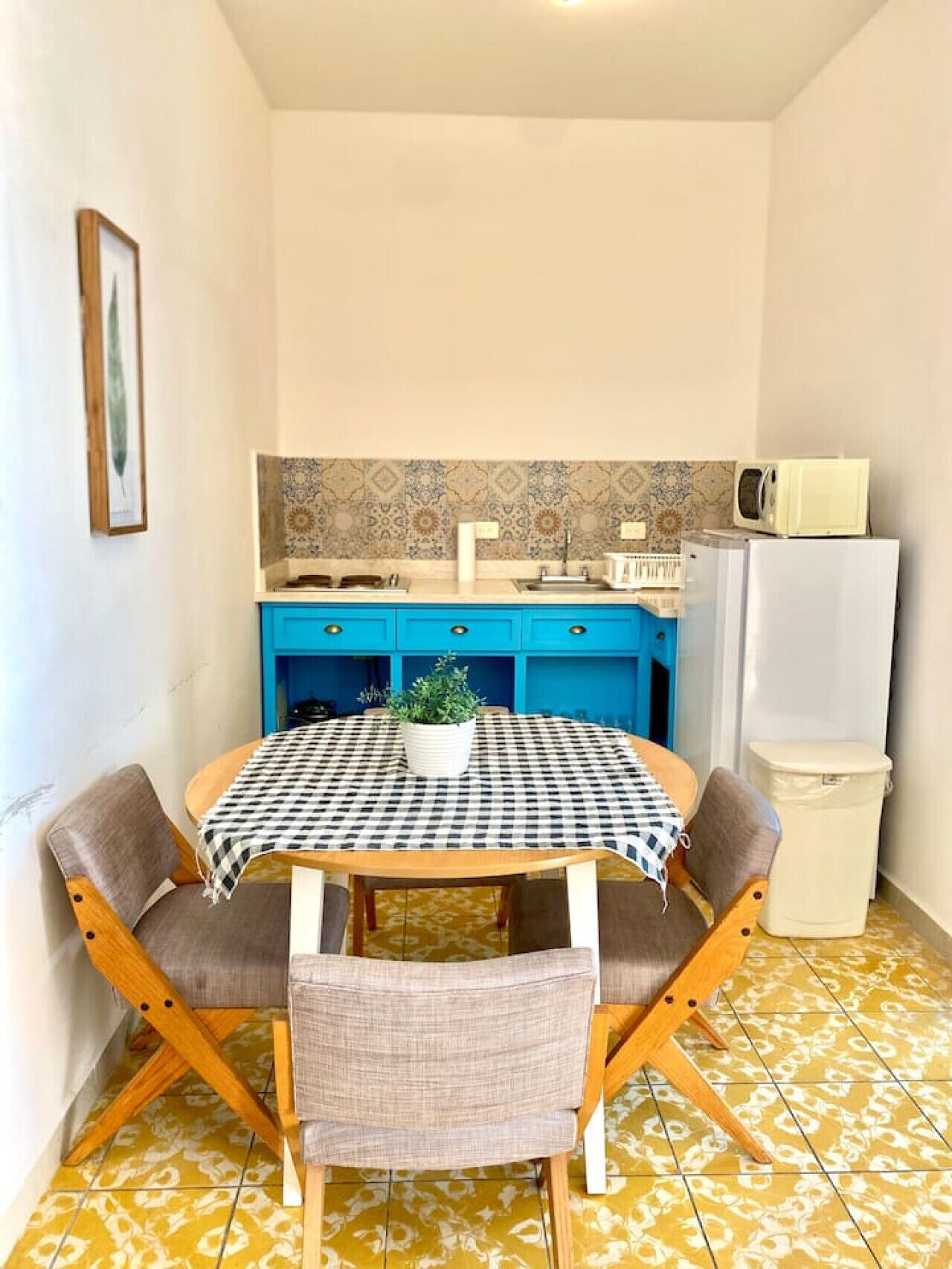 Una cocina compacta con armarios azules, un microondas y un refrigerador. En primer plano, una mesa a cuadros con sillas.