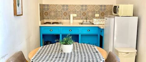 Una cocina compacta con armarios azules, un microondas y un refrigerador. En primer plano, una mesa a cuadros con sillas.