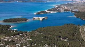 An aerial view of a coastal area with a fortified structure surrounded by water, dense greenery, and a distant town. - Apartment in Brodarica Near Beach Gaj (Sibenik)