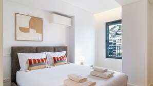 A modern hotel room with a large bed, white linens, a grey headboard, and a window with a city view. - GuestReady - Acqua residences - Aquamarine (Funchal)