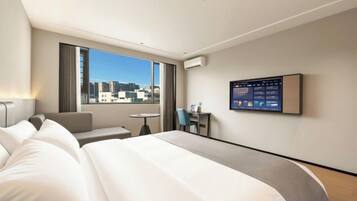 Habitación de hotel con una cama grande, un escritorio, una silla, una mesita y un ventanal con vistas a la ciudad.