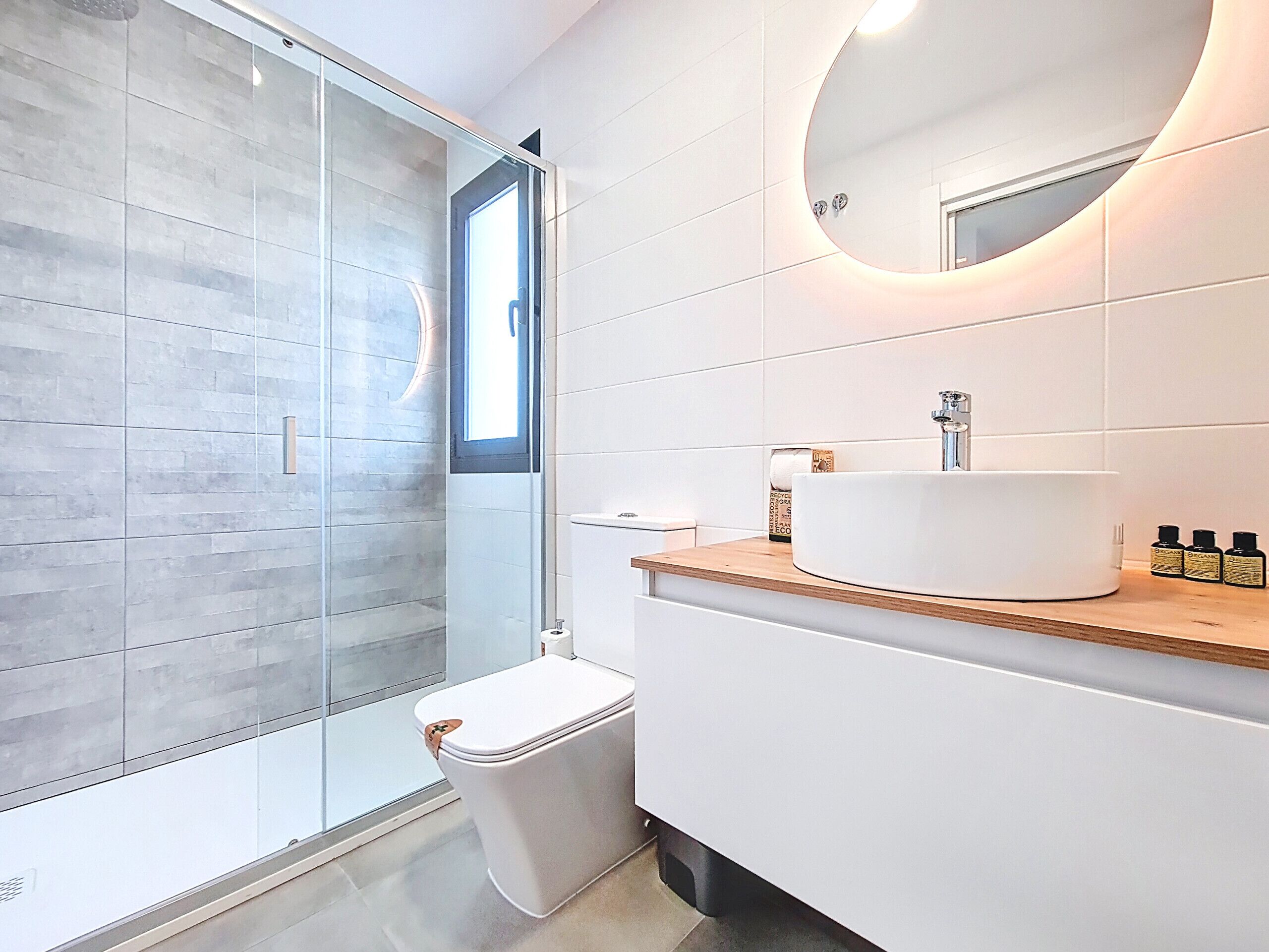 A modern bathroom with a glass shower enclosure, a white toilet, a round sink with a wooden countertop, and a mirror with a light above it.