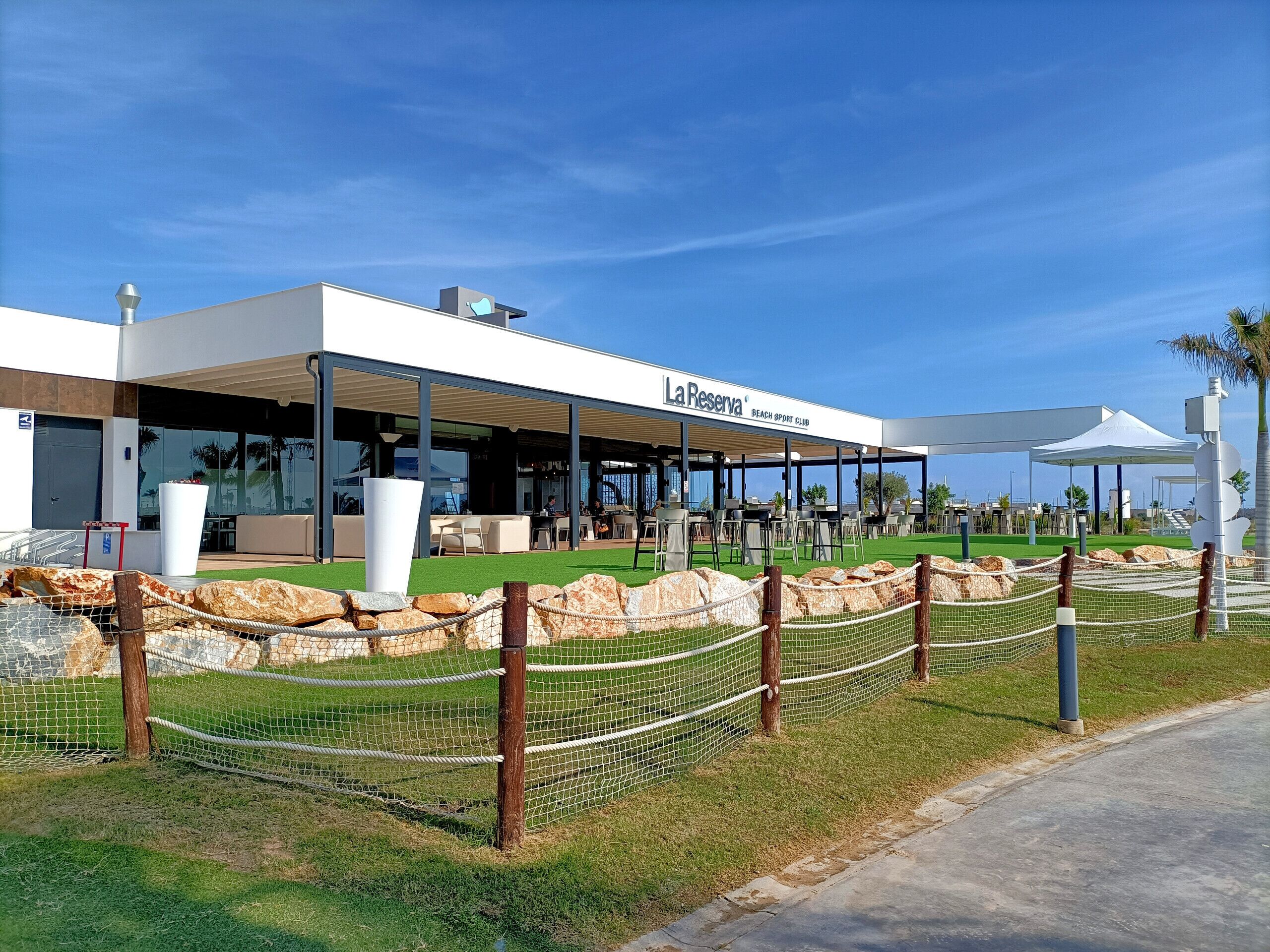 A modern building with a covered outdoor seating area, a sign reading "La Reserva Beach Sport Club," and a fenced area with rocks and grass.