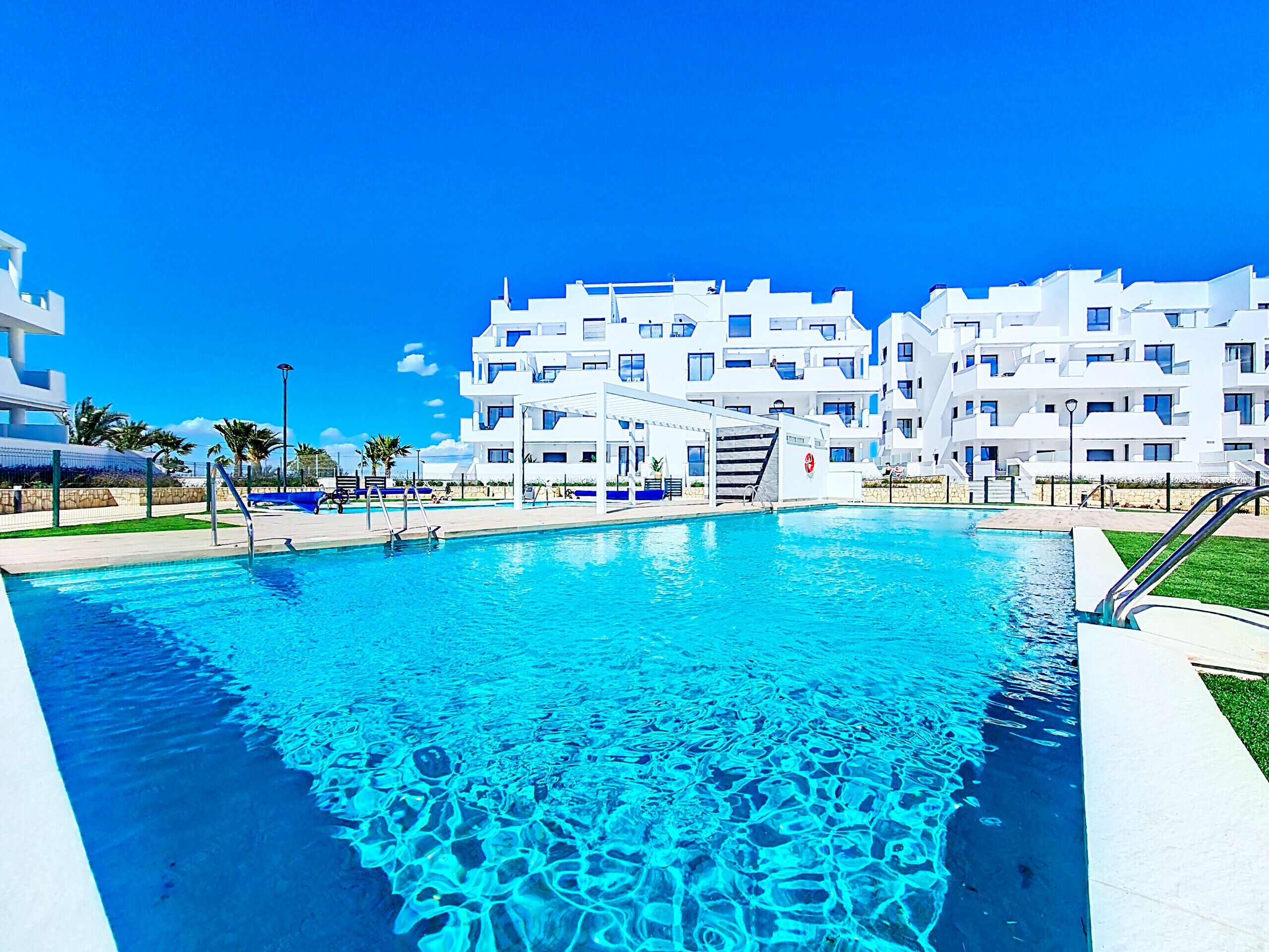 A swimming pool with clear blue water, surrounded by white buildings with balconies, and a clear blue sky.