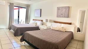 Une chambre d’hôtel avec deux lits, une grande fenêtre avec des rideaux, un ventilateur de plafond et des œuvres d’art accrochées aux murs.