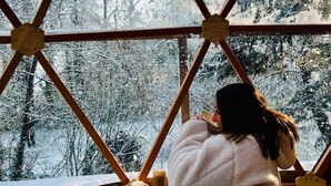 A room with a large window featuring a geometric wooden frame, a bed with a blanket, and a candle on the windowsill.