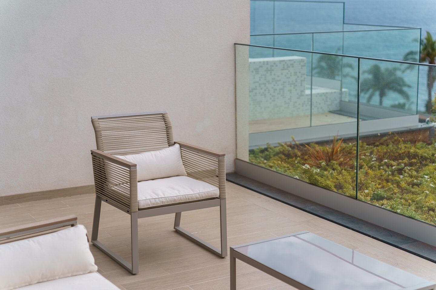A modern lounge area with a wicker chair and a glass table, overlooking a landscaped garden and a building with a glass facade.