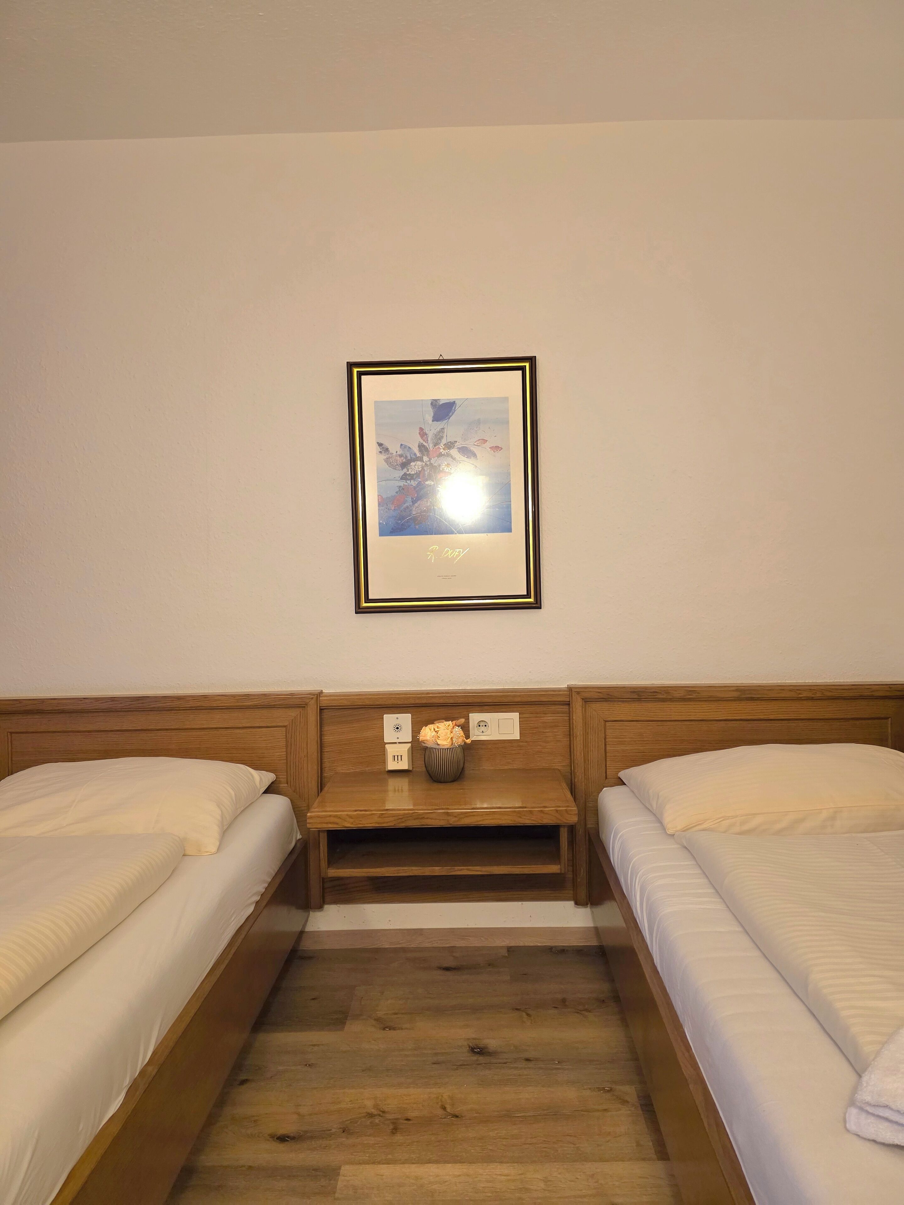Habitación de hotel con dos camas, cabecero de madera, una mesita con un florero y un cuadro en la pared.