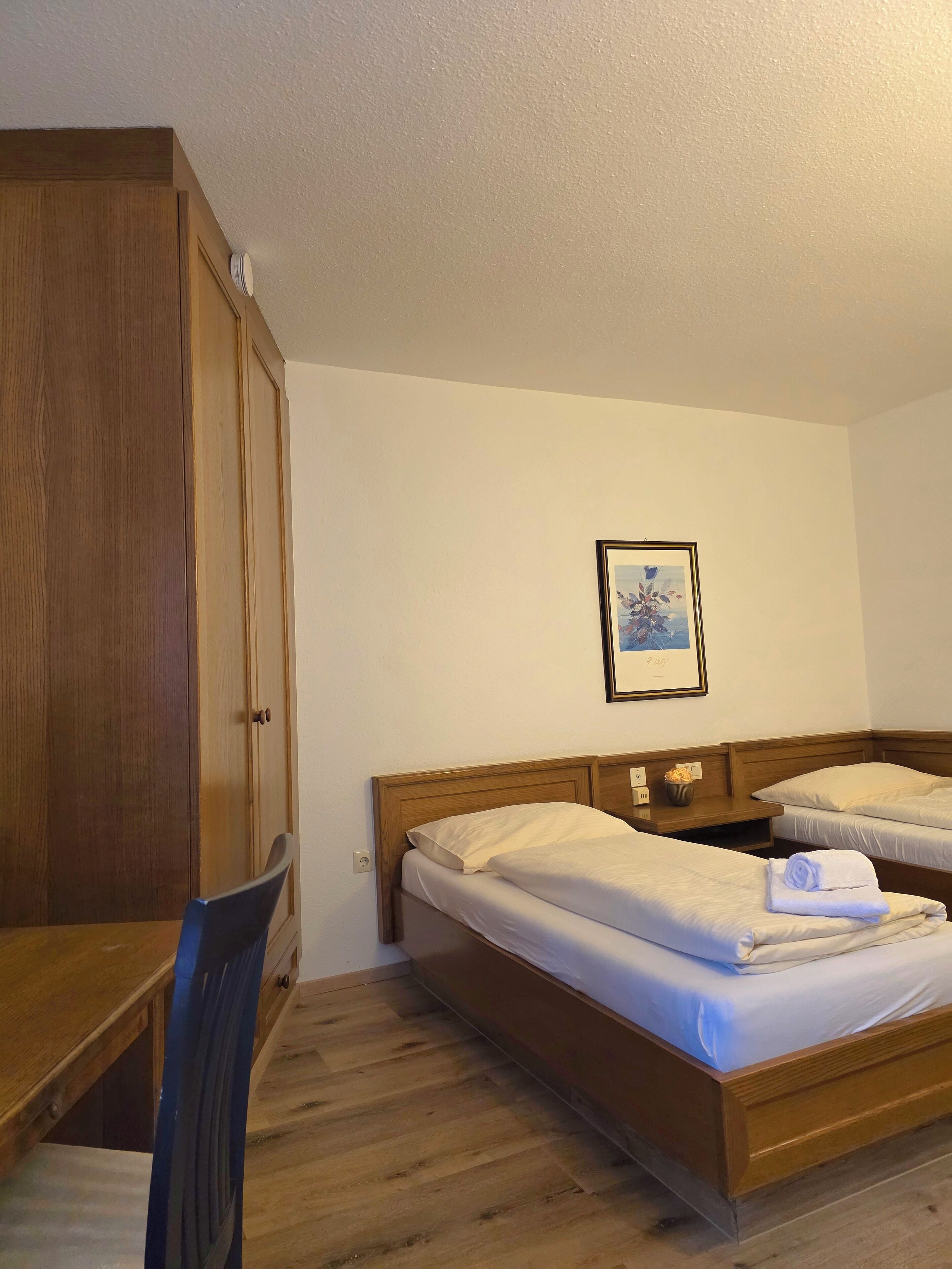 A hotel room with two single beds, a wooden desk, a chair, and a framed picture on the wall.